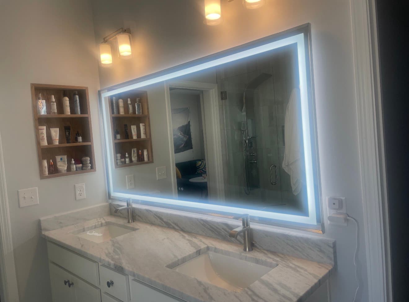 Double vanity bathroom with a large backlit mirror, marble-look counter, and recessed wood storage niches.