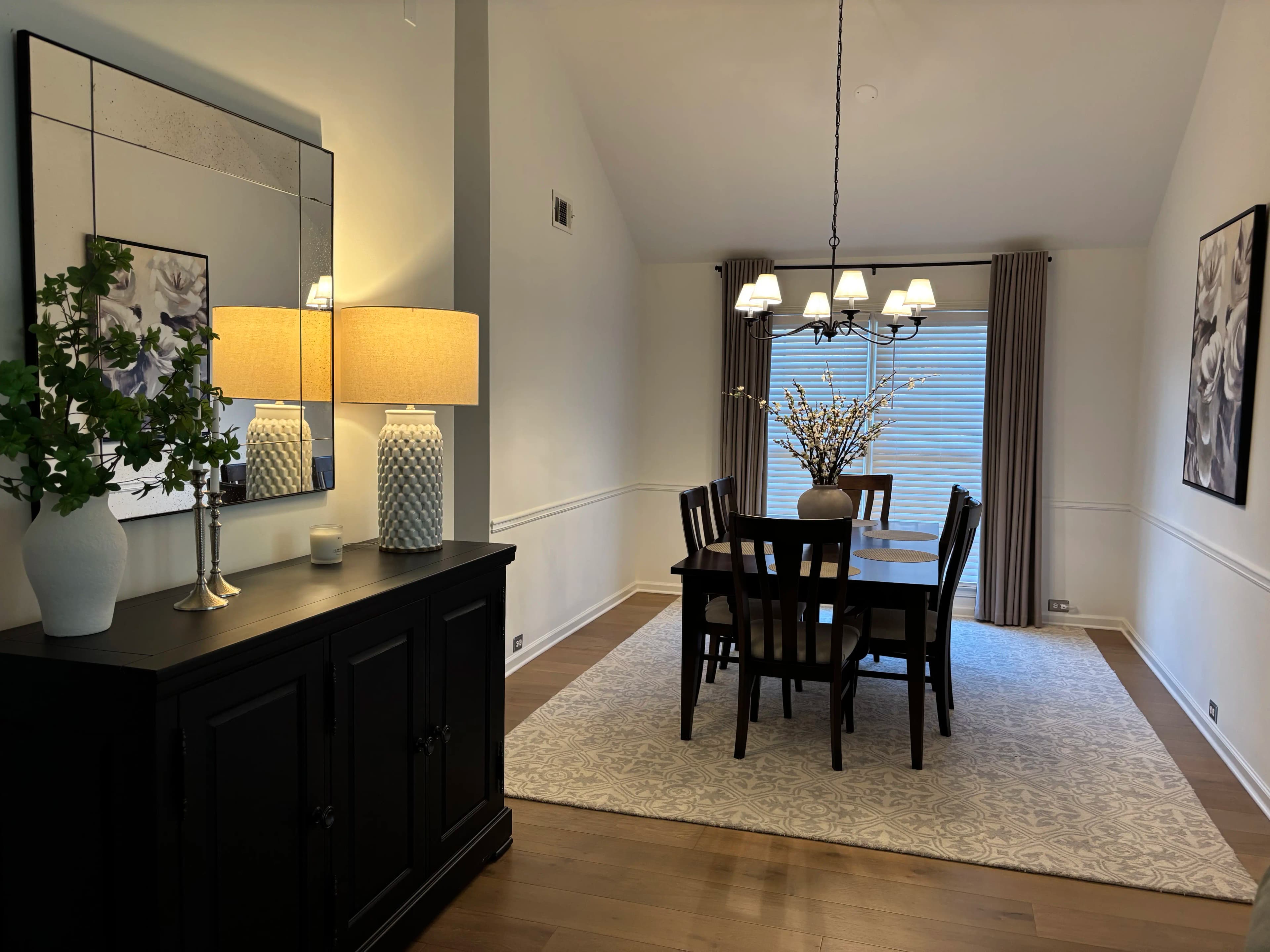 Formal dining room with a dark sideboard, patterned mirror, lamps, drapery, and a six-seat dining table under a chandelier.