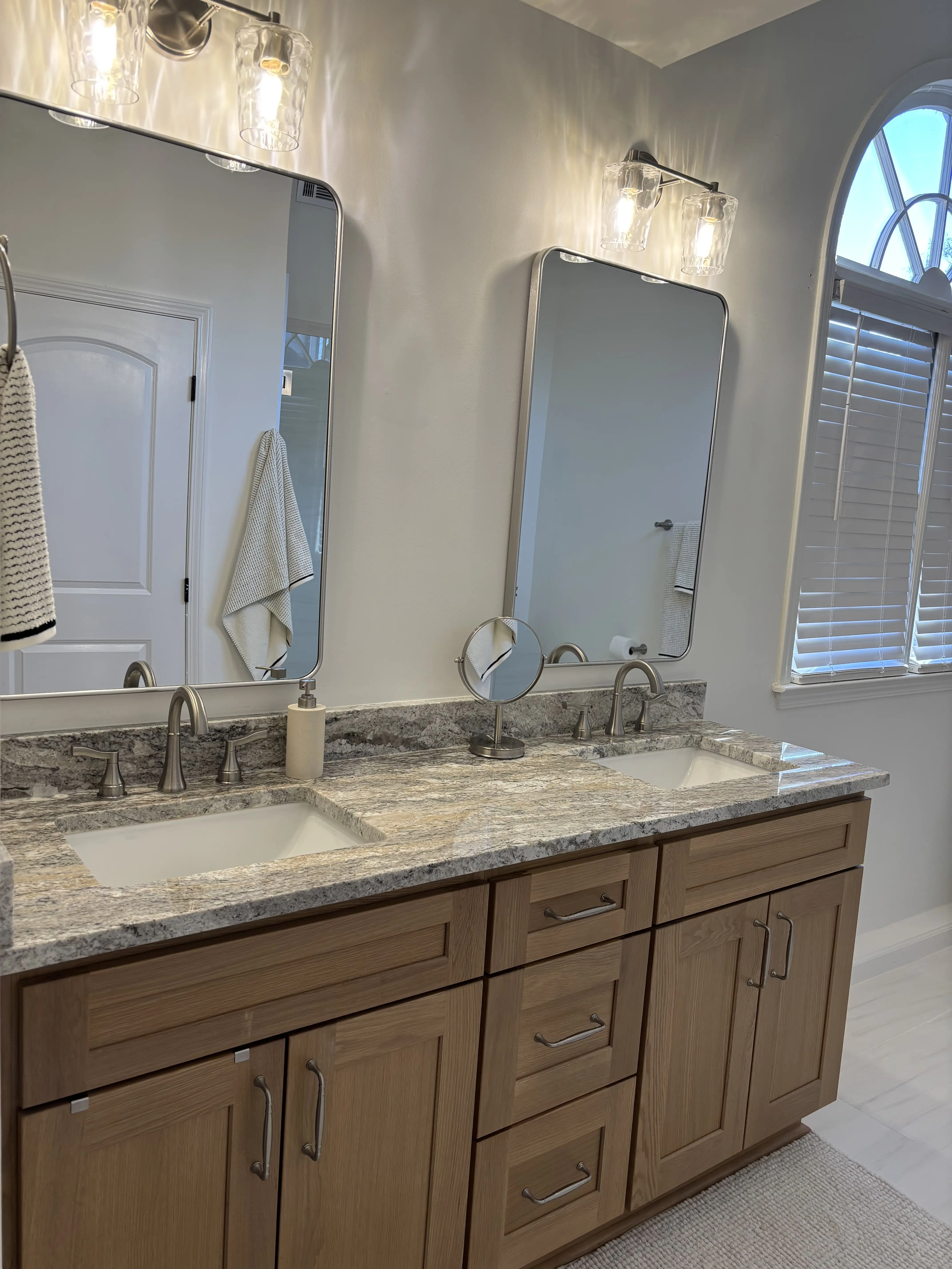 Primary bathroom remodel with a warm wood double vanity, stone countertop, two tall mirrors, and polished wall sconces.